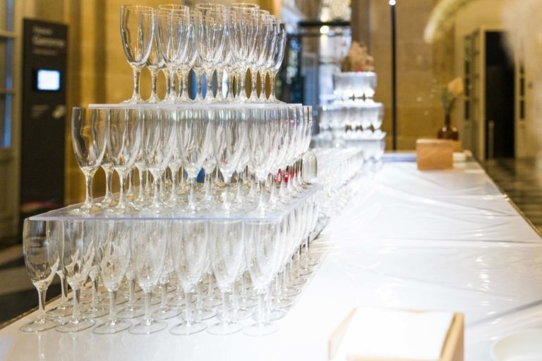 Une pyramide de flûtes à champagne vides est exposée sur une longue table blanche lors de la Soirée AXA Business dans l'élégant Palais de la Bourse de Bordeaux. Les verres sont soigneusement empilés en rangées, créant une structure à plusieurs niveaux. L'arrière-plan présente un espace intérieur doucement éclairé avec une grande plante à droite et un bloc de bois sur la table.