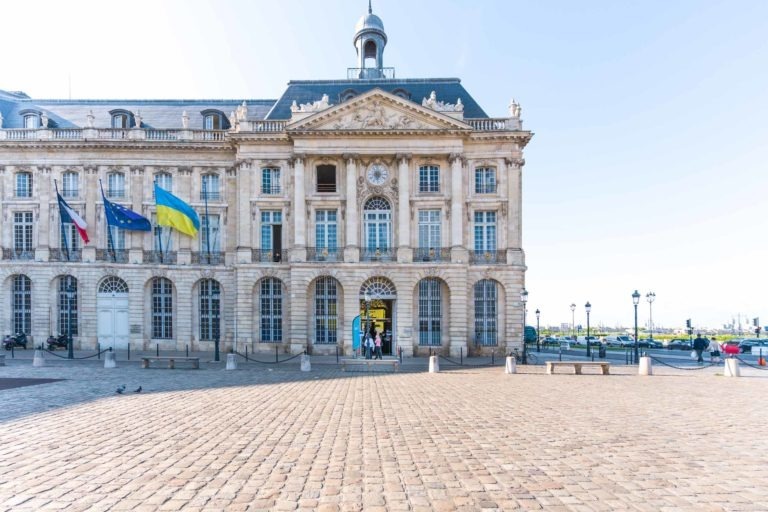 Un grand bâtiment historique avec des arcades et une horloge au-dessus de son entrée principale, situé sur une place pavée. Des drapeaux français et ukrainiens flottent sur le côté gauche du Palais de la Bourse de Bordeaux. On peut voir des gens entrer et sortir par les portes pour une soirée. Le ciel est clair et bleu.