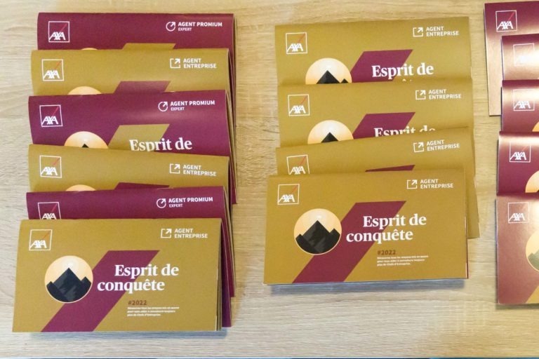 Image montrant deux piles de livrets AXA posées sur une surface en bois. À gauche, une pile de livrets rouges intitulés « Esprit de conquête » avec un logo de montagne noir et « 2022 » en dessous. La pile de droite présente des livrets à couverture jaune-orange avec le même design et le même titre, provenant de la Soirée AXA Business au Palais de la Bourse de Bordeaux.