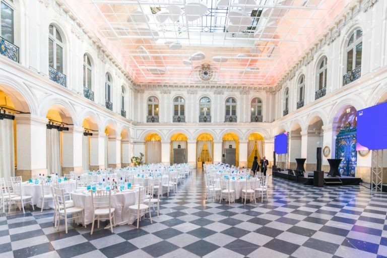 Salle de réception élégante avec de hauts plafonds et des fenêtres cintrées, avec un sol à carreaux noir et blanc. De nombreuses tables rondes sont soigneusement disposées avec des nappes blanches et des couverts. Une scène avec deux grands écrans et des podiums se trouve sur le côté droit, alors que les gens se préparent pour la Soirée AXA au Palais de la Bourse de Bordeaux.