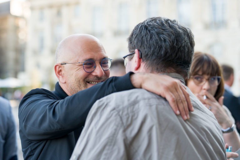 Deux hommes interagissent dans un décor extérieur lors de la Soirée AXA Business. L'homme de gauche, chauve avec des lunettes, portant une veste noire, sourit et passe son bras autour de l'autre homme. Le deuxième homme, aux cheveux gris et portant des lunettes, porte une veste gris clair. En arrière-plan au Palais de la Bourse de Bordeaux, une femme tient une cigarette.