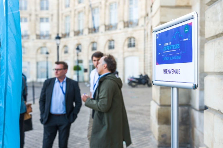 Plusieurs personnes en tenue professionnelle discutent à l'extérieur, près d'un bâtiment en pierre. Un panneau sur la droite indique « Bienvenue » et porte l'en-tête « Expériences Edition Sud-Ouest ». L'arrière-plan montre une cour avec une moto garée à proximité, des détails architecturaux classiques et des allusions à la prochaine Soirée AXA Business au Palais de la Bourse de Bordeaux.