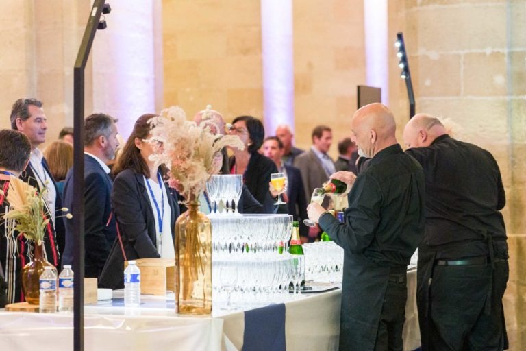 Des gens discutent lors d'une soirée en salle au AXA Business Palais de la Bourse de Bordeaux. Deux barmans servent des boissons à une longue table décorée de verres en cristal et de compositions florales dans de grands vases. Les serveurs en tenue de soirée discutent tout en tenant leurs boissons. Un éclairage chaleureux renforce l'ambiance élégante.