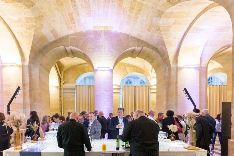 Les invités se réunissent dans une élégante salle voûtée aux murs de pierre et aux hauts plafonds pour la soirée AXA Business au Palais de la Bourse à Bordeaux. Ils discutent et dégustent des boissons. Un bar bien éclairé est installé au centre avec deux barmans servant des boissons, orné de compositions florales et de bouteilles de vin, renforçant l'atmosphère festive.