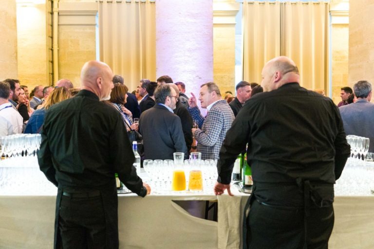 Lors de la soirée AXA Business au Palais de la Bourse de Bordeaux, un large public est réuni dans une salle bien éclairée. Deux serveurs en uniformes noirs se tiennent derrière une table remplie de verres et de bouteilles. La table propose également du jus d'orange et un autre pichet contenant un liquide jaune, encadrés par des rideaux beiges éclairés par une lumière chaude.