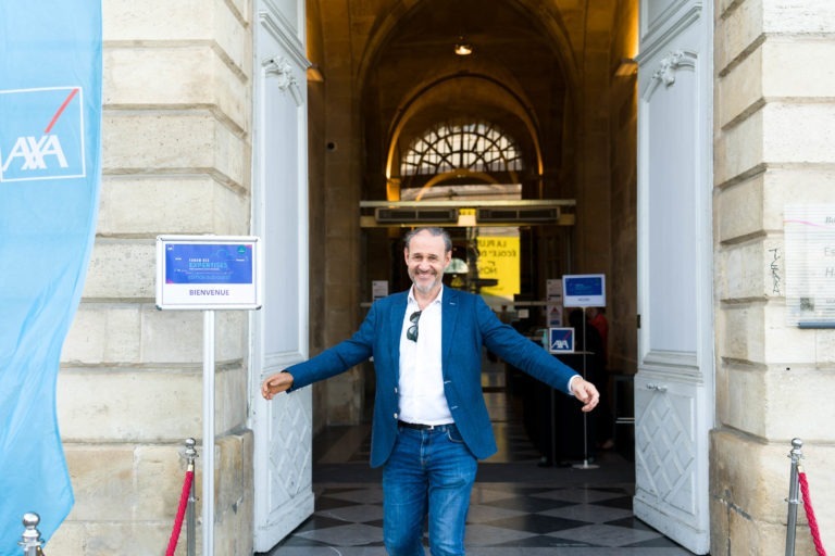 Un homme en blazer bleu et chemise blanche, lunettes de soleil à la main, accueille les visiteurs à bras ouverts à l'entrée d'un bâtiment Axa. Les éléments architecturaux imposants, comme les murs en pierre et les portes cintrées, préparent le terrain pour la Soirée AXA Business au Palais de la Bourse de Bordeaux. À sa gauche, un panneau avec "BIENVENUE" et le logo Axa est visible.