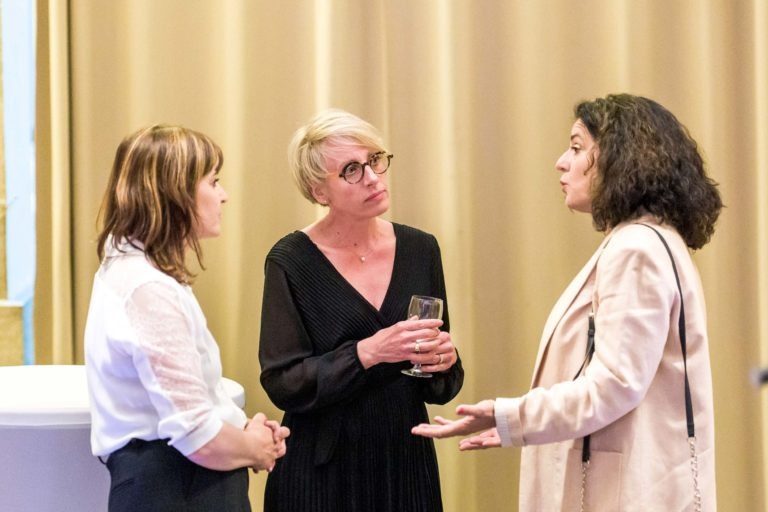 Lors de la Soirée AXA Business, trois femmes discutent devant un rideau doré au Palais de la Bourse de Bordeaux. La femme de gauche porte un chemisier blanc, celle du milieu aux cheveux blonds courts porte des lunettes et une robe noire tout en tenant un verre, et la femme de droite arbore des cheveux noirs bouclés et un manteau clair.