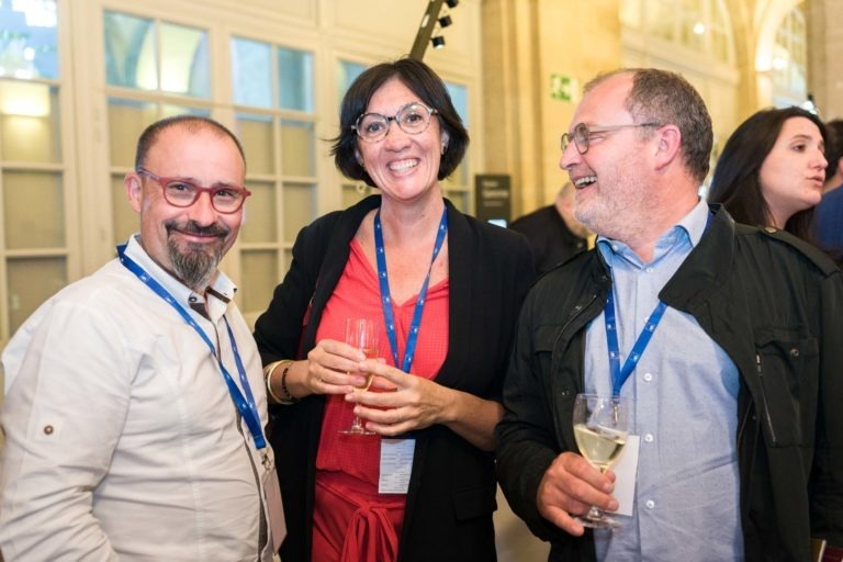 Trois personnes, deux hommes et une femme, se tiennent debout et sourient lors de la Soirée AXA Business au Palais de la Bourse de Bordeaux. Ils ont tous un cordon autour du cou et tiennent un verre de vin blanc. La femme au centre porte une tenue rouge, tandis que les hommes sont habillés de chemises claires. Ils semblent passer un bon moment