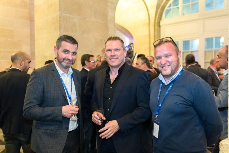 Trois hommes en tenue professionnelle se tiennent ensemble lors d'une soirée en salle au Palais de la Bourse à Bordeaux, souriant à la caméra. L'homme de gauche tient un verre, celui du milieu porte un costume sombre et celui de droite a des lunettes sur la tête. D'autres invités se mêlent à l'arrière-plan.