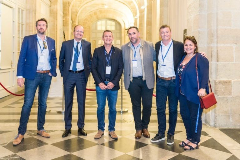 Un groupe de six personnes, quatre hommes et deux femmes, se tiennent dans un couloir avec des arches et un sol en damier lors de la Soirée AXA Business au Palais de la Bourse de Bordeaux. Ils sont habillés en tenue professionnelle décontractée, portent des badges de conférence et certains sourient, les mains dans les poches ou se prennent dans les bras. Le cadre semble formel et élégant.