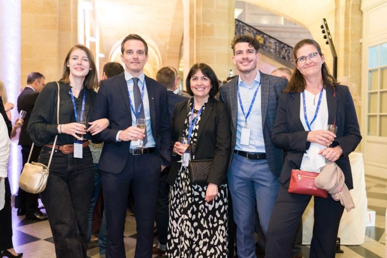 Cinq personnes se tiennent debout, souriantes, en tenant un verre à la main lors de la Soirée AXA Business dans le grand hall du Palais de la Bourse de Bordeaux aux murs en pierre. Trois d'entre elles portent un costume, tandis que les deux autres sont en tenue professionnelle. Elles ont toutes un cordon autour du cou et semblent heureuses. D'autres participants et un escalier sont visibles en arrière-plan.