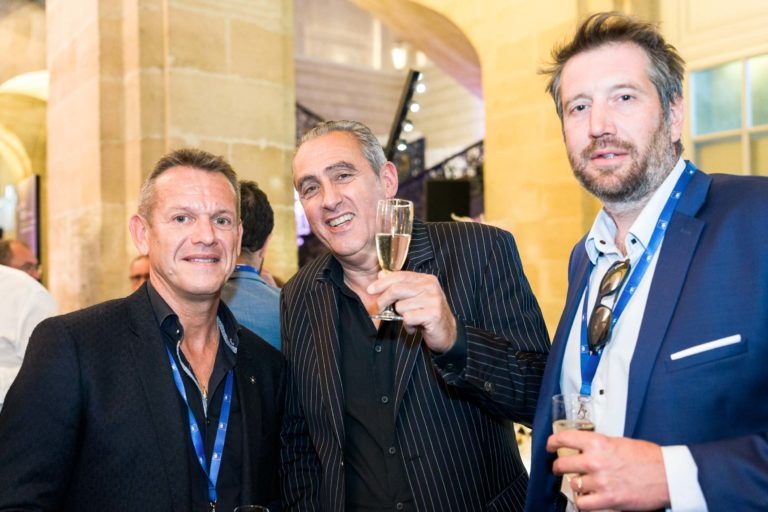 Trois hommes, habillés en tenue professionnelle, se tiennent ensemble dans un espace intérieur bien éclairé lors de la Soirée AXA. L'homme de gauche porte un costume sombre et un cordon, tandis que l'homme du milieu, également en tenue sombre, lève un verre de champagne. L'homme de droite, vêtu d'une veste bleue et d'une chemise blanche, tient un verre au Palais de la Bourse de Bordeaux.