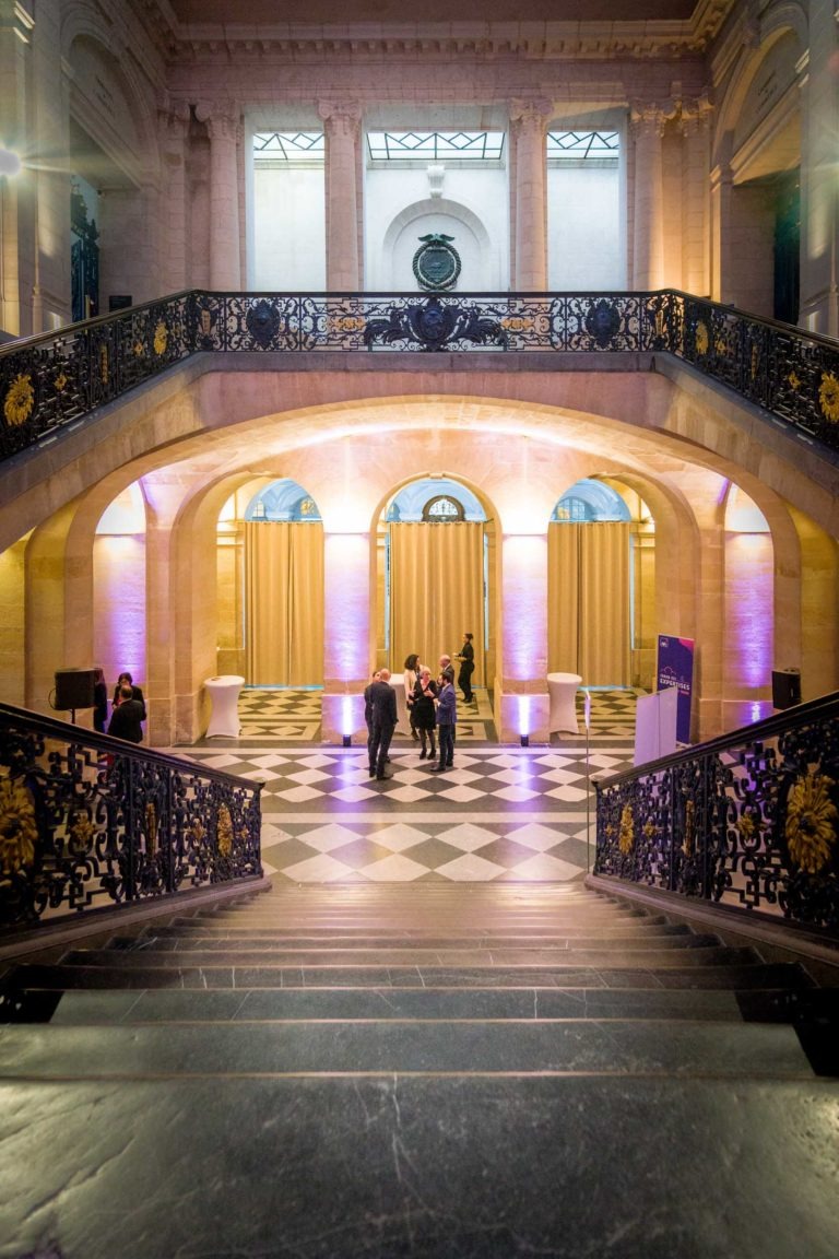 Un intérieur élégant au Palais de la Bourse de Bordeaux avec de grands escaliers en pierre ornés de rampes noires et dorées complexes. L'espace, parfait pour une soirée AXA Business, présente un sol à carreaux noir et blanc et de hautes colonnes arquées illuminées par une lumière violette. De hauts plafonds et de grandes fenêtres rehaussent la grandeur tandis que des groupes de personnes discutent au centre.