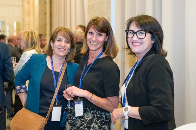 Trois femmes se tiennent côte à côte, souriantes et posent pour une photo lors d'une soirée au Palais de la Bourse à Bordeaux. Elles sont habillées en tenue de ville décontractée, avec des cordons et des badges nominatifs visibles. Deux d'entre elles tiennent des verres à vin. L'arrière-plan montre d'autres participants et une salle aux rideaux beiges.