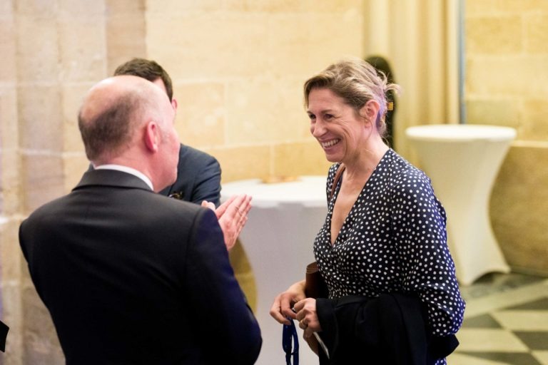 Lors d'une soirée au Palais de la Bourse de Bordeaux, une femme en robe à pois noirs et blancs sourit et discute avec un homme chauve en costume sombre. Ils se tiennent devant un mur de pierre avec une table haute blanche en arrière-plan. L'homme, les mains levées, parle avec animation lors de cette réunion d'affaires d'AXA.