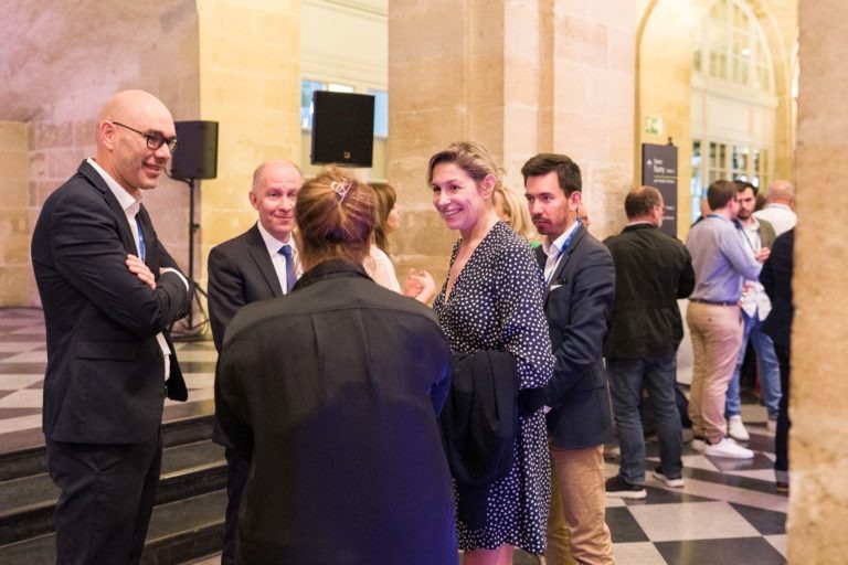 Un groupe de personnes discutent dans une salle élégante et chaleureuse aux murs en pierre et au sol à carreaux lors de la Soirée AXA Business qui s'est tenue au Palais de la Bourse à Bordeaux. Au premier plan, deux hommes en costume-cravate et une femme en robe à pois noirs et blancs discutent. D'autres personnes discutent à l'arrière-plan.