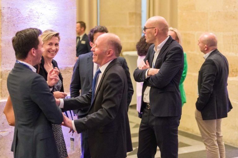 Un groupe de sept personnes en tenue professionnelle discutent et rient lors d'un événement en intérieur. Trois hommes font face à une femme qui sourit, tandis que deux autres hommes ont le dos tourné. Des murs en pierre et un éclairage tamisé créent une atmosphère à la fois professionnelle et décontractée lors de la soirée AXA Business qui se tient au Palais de la Bourse de Bordeaux.