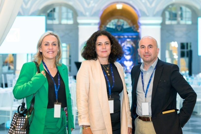 Trois personnes se tiennent à l'intérieur de l'événement Soirée AXA Business, habillées en tenue décontractée. La personne à gauche porte un costume vert, celle du milieu un blazer beige clair et celle de droite un blazer bleu marine. Elles portent toutes des badges de conférence et sourient devant un intérieur élégant avec des portes cintrées et un éclairage tamisé.