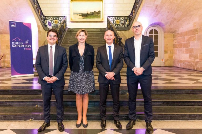 Quatre personnes se tiennent dans un grand couloir au sol carrelé à carreaux et derrière elles se trouve un large escalier au Palais de la Bourse de Bordeaux. À gauche, une bannière violette sur laquelle est écrit « France des Expertises ». Les individus sont habillés en tenue de travail formelle, peut-être ici pour la Soirée AXA Business. Un grand tableau est visible en haut de l'escalier.