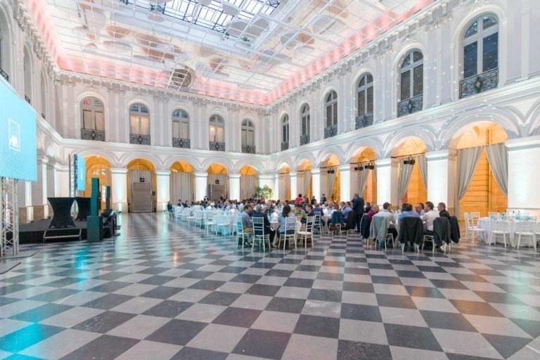 Une grande salle élégante, aux hauts plafonds et aux détails architecturaux complexes, est remplie de personnes assises à des tables rondes. Des lustres ornent le plafond et un toit en verre permet à la lumière naturelle d'illuminer l'espace. L'atmosphère grandiose est parfaite pour la Soirée AXA Business au Palais de la Bourse de Bordeaux.