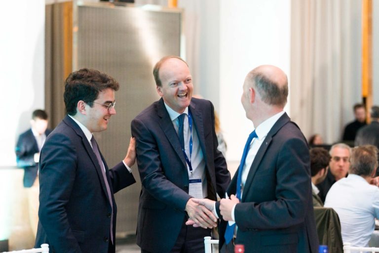 Lors de la soirée AXA Business qui s'est tenue au grand Palais de la Bourse à Bordeaux, trois hommes en costume échangent chaleureusement. L'homme du milieu sourit largement et serre la main de l'homme de droite, tandis que l'homme de gauche a la main sur l'épaule. L'arrière-plan révèle d'autres convives et un décor flou.