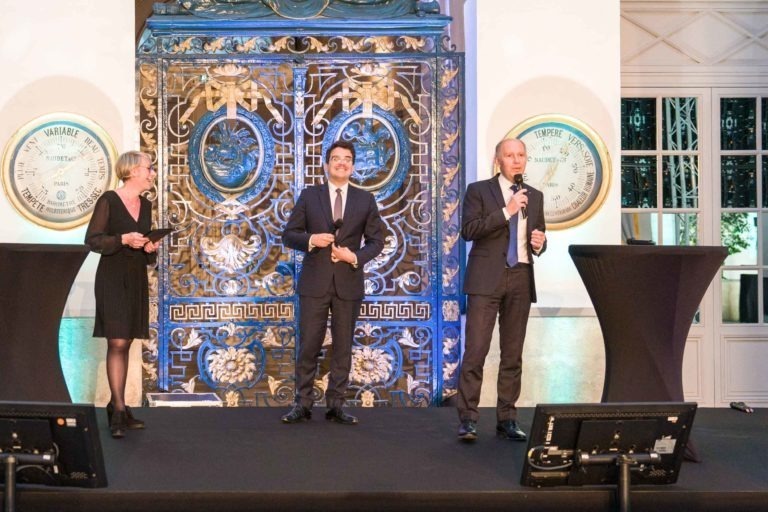 Trois personnes se tiennent sur une scène lors de la Soirée AXA Business au Palais de la Bourse de Bordeaux. Deux hommes en costume-cravate tiennent des micros et parlent, tandis qu'une femme en robe noire écoute à gauche. Derrière eux se trouve un décor orné de motifs complexes et d'horloges affichant « Variable » et « Tempête ». Des tables recouvertes de nappes noires encadrent la scène.