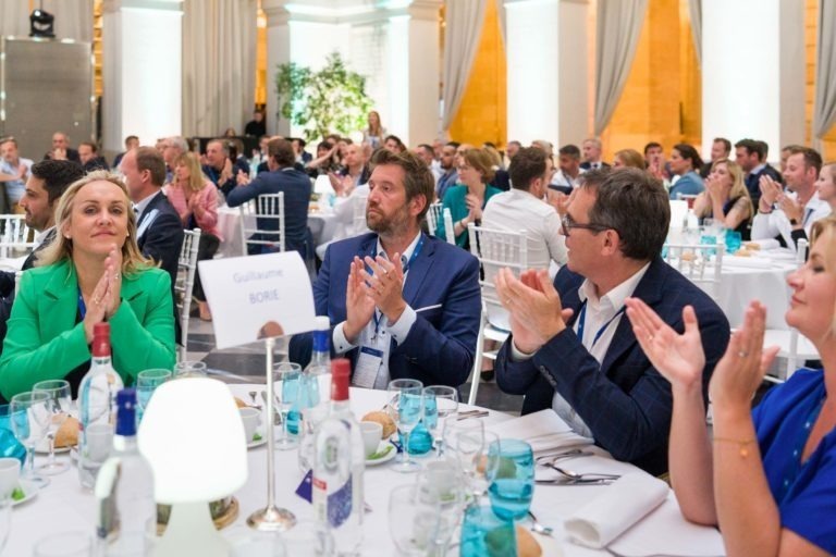 Un grand groupe de personnes est assis à des tables rondes dans une élégante salle de banquet lors de la Soirée AXA Business au Palais de la Bourse à Bordeaux. Ils applaudissent et regardent vers l'avant. La table au premier plan porte une pancarte avec le nom de « Guillaume Borie ». Les participants sont bien habillés et la table est dressée avec des verres, du pain et une table blanche