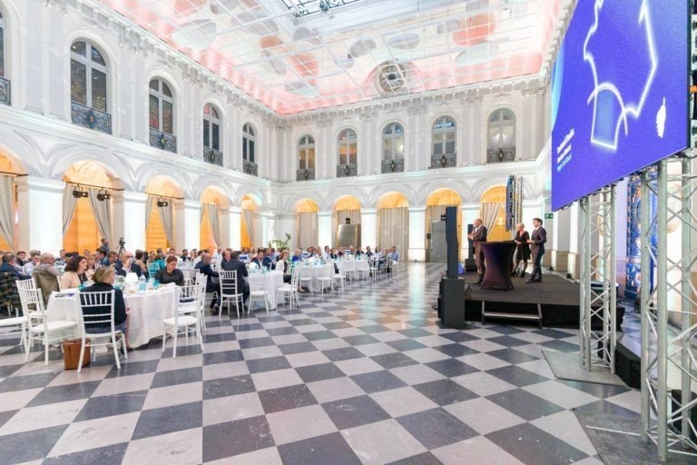 La Soirée AXA Business se déroule dans une élégante salle de réception au Palais de la Bourse de Bordeaux, avec de hauts plafonds et de grandes fenêtres cintrées. Les participants sont assis à des tables rondes avec des chaises et des nappes blanches. Un orateur sur scène s’adresse au public à côté d’un grand écran diffusant une présentation bleue, encadrée par le sol à carreaux noirs et blancs de la salle.