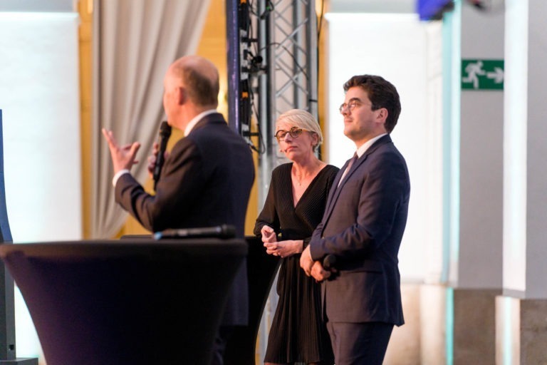 Trois personnes en tenue de soirée sont sur scène lors de la Soirée AXA Business au Palais de la Bourse de Bordeaux. Une personne parle dans un micro, dos à la caméra, tandis qu'une femme et un homme regardent vers l'avant et écoutent attentivement. La salle est bien éclairée, de hauts piliers et un panneau de sortie est visible.