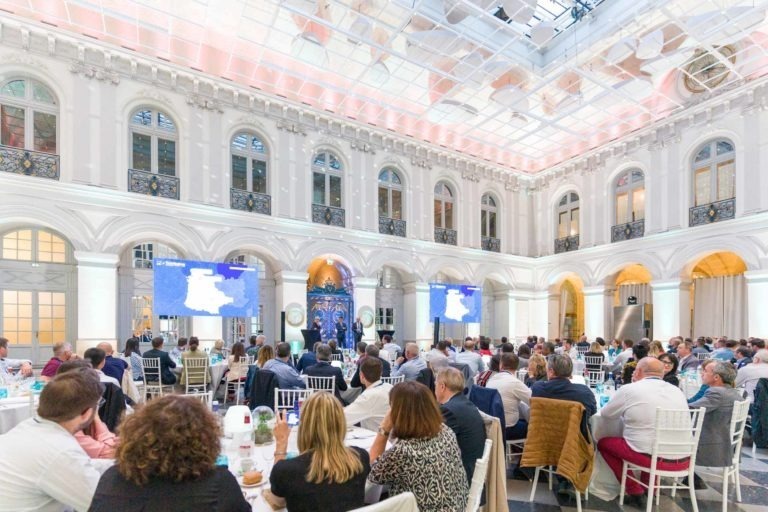 Lors de la Soirée AXA Business qui se tient au Palais de la Bourse de Bordeaux, un large groupe de personnes est assis autour de tables rondes dans un espace événementiel au design raffiné, haut de plafond, avec des fenêtres cintrées et un toit en verre. Deux écrans à l'avant diffusent une présentation, tandis qu'un éclairage blanc doux met en valeur son décor et son architecture élégants.