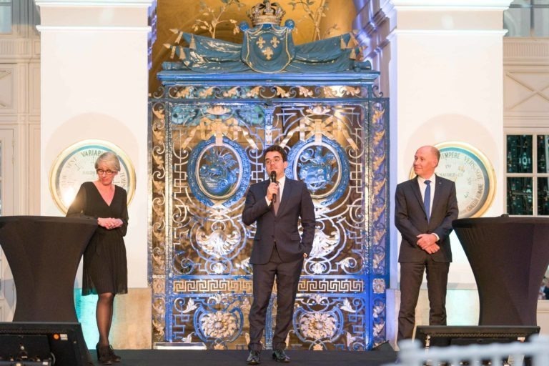 Trois personnes en tenue de soirée se tiennent sur une scène décorée de façon raffinée lors de la Soirée AXA Business qui se tient au Palais de la Bourse, à Bordeaux. La personne au centre parle dans un micro, tandis que les autres se tiennent de chaque côté derrière des podiums. Le décor présente des motifs complexes bleus et dorés dans cette grande salle aux hauts plafonds.