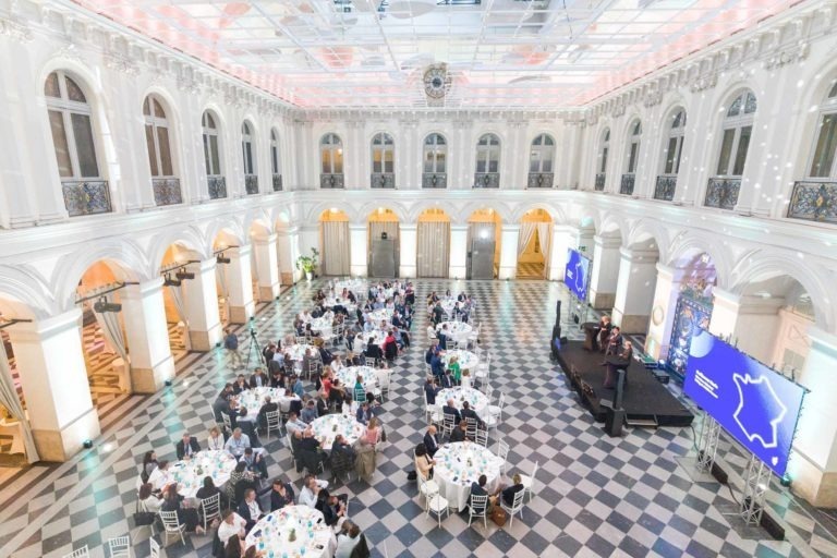 Une grande salle de réception élégante avec un sol en damier et de hauts murs voûtés illuminés de blanc. Plusieurs tables rondes avec des nappes blanches et des invités assis sont répartis dans la salle. Lors de la Soirée AXA Business qui se tient au Palais de la Bourse à Bordeaux, les participants dînent et discutent tandis qu'une scène à l'avant est équipée de haut-parleurs et de grands écrans affichant un contour de carte.