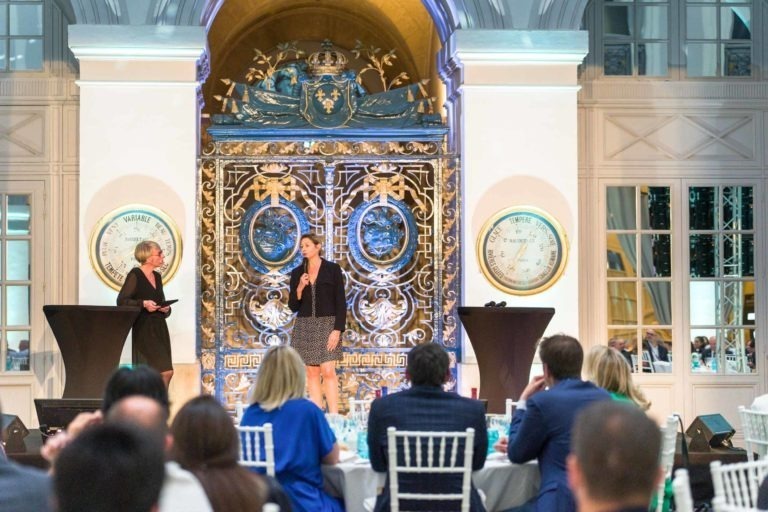 Lors de la Soirée AXA Business au Palais de la Bourse de Bordeaux, un orateur se tient sur scène avec un micro et s'adresse à un public assis à des tables blanches. Une autre personne se tient sur le côté. La scène est ornée d'un fond bleu et doré avec de grands médaillons, d'un plafond voûté doré et de deux grandes horloges.