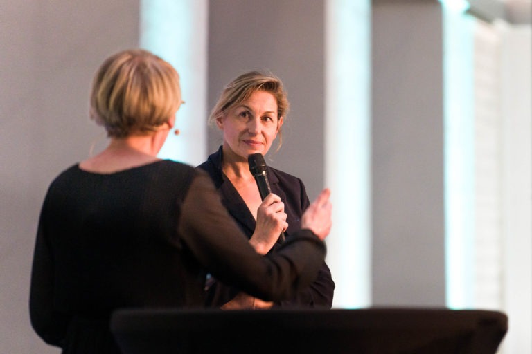 Deux femmes discutent lors de la Soirée AXA Business qui se tient au Palais de la Bourse de Bordeaux. La femme de droite, vêtue d'un blazer noir, tient un micro et a l'air attentive. La femme de gauche, aux cheveux blonds courts et tenant également un micro, fait des gestes animés. L'arrière-plan est doucement éclairé par un ton neutre.