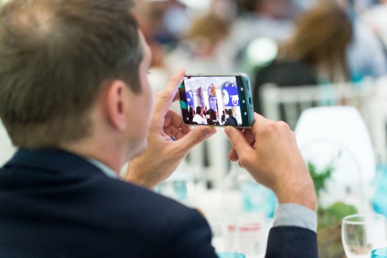 Un homme en costume tient un smartphone à l'horizontale, prenant une photo ou une vidéo d'un événement. L'écran affiche une scène avec un intervenant et une bannière de la Soirée AXA Business. L'arrière-plan est flou avec des personnes assises à des tables, suggérant une conférence ou un rassemblement officiel au Palais de la Bourse de Bordeaux.