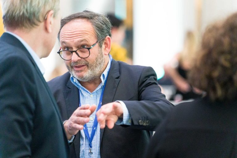 Lors de la Soirée AXA Business au Palais de la Bourse de Bordeaux, un homme portant des lunettes et une barbe, vêtu d'un costume et d'une chemise à carreaux bleus, engage la conversation. Il fait des gestes appuyés tandis qu'une autre personne aux cheveux blonds, également en costume, l'écoute attentivement. Des personnages flous et une pièce très éclairée forment l'arrière-plan.