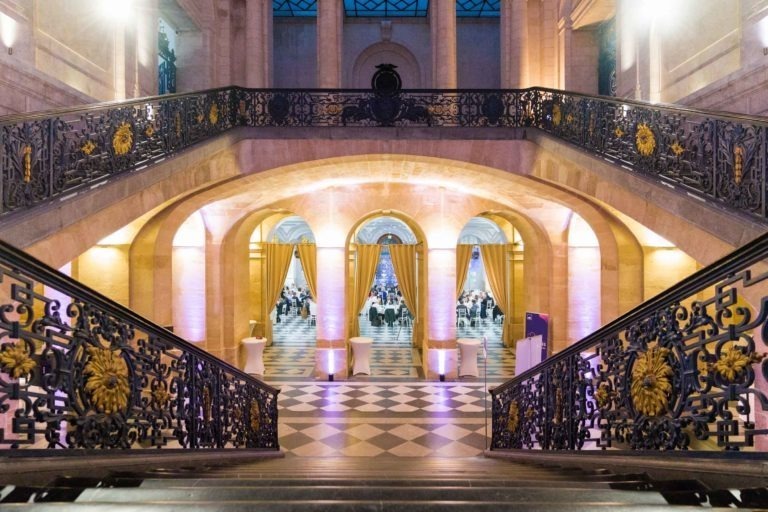 Un escalier décoré de décorations dorées mène à un grand hall aux portes cintrées drapées de rideaux jaunes. Le hall, parfait pour une soirée à l'AXA Business Palais de la Bourse de Bordeaux, présente un sol à carreaux noirs et blancs et un éclairage doux et doré, créant une ambiance élégante.