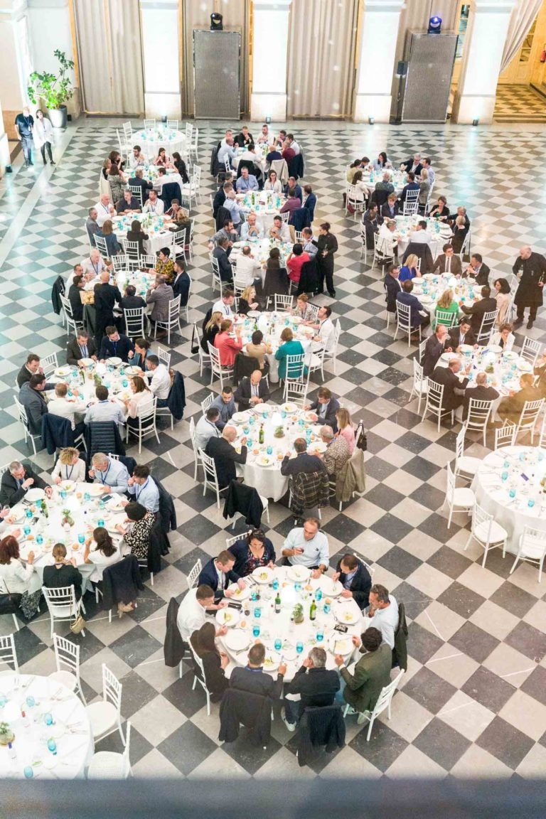 Une grande et élégante salle de bal au Palais de la Bourse de Bordeaux, au sol à carreaux noirs et blancs, abrite plusieurs tables rondes, chacune entourée de personnes en conversation. Les tables sont ornées de verres bleus et de centres de table. Un éclairage vif provenant de hautes fenêtres illumine la salle, renforçant son ambiance de soirée festive.