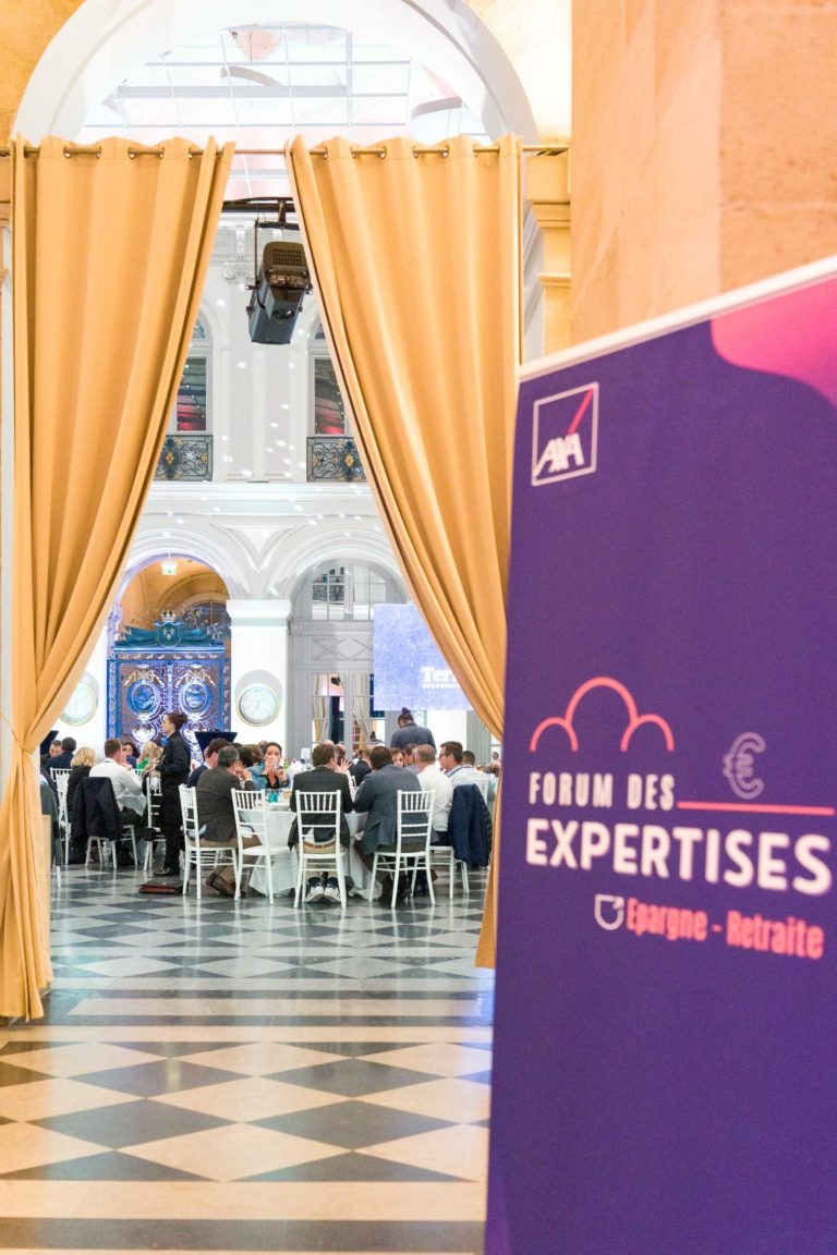 Une salle de conférence chaleureusement éclairée avec un sol à carreaux noirs et blancs, ornée de hauts rideaux jaunes. A l'intérieur, les gens sont assis à des tables rondes, discutant. Au premier plan, une banderole violette indique "Soirée AXA Business" avec des thèmes financiers sur la retraite et l'épargne au Palais de la Bourse de Bordeaux.