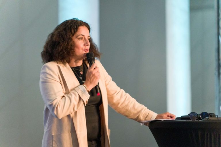 Une femme aux cheveux bouclés mi-longs se tient derrière une table ronde lors de la Soirée AXA Business au Palais de la Bourse de Bordeaux, parlant dans un micro à main. Elle porte un blazer beige clair sur une chemise noire. L'arrière-plan est flou, avec de hauts piliers blancs et un éclairage doux. Une paire de lunettes repose sur la table à côté d'elle.