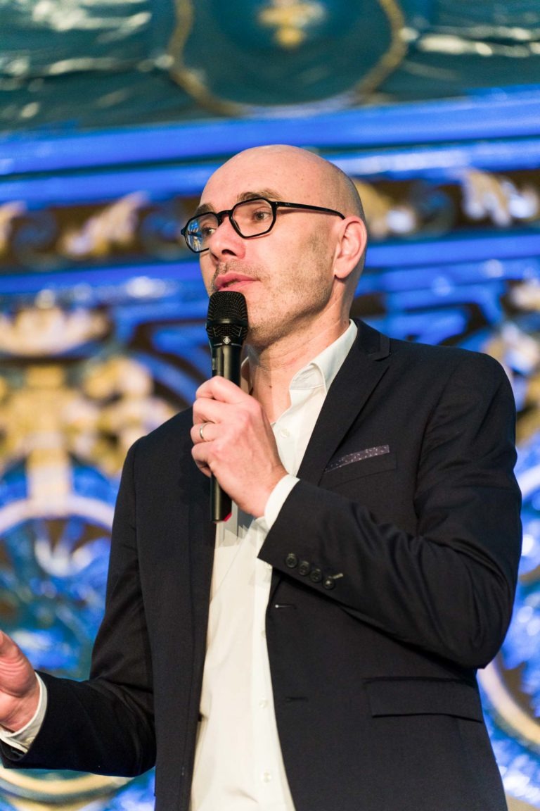 Un homme chauve portant des lunettes, une chemise blanche et un blazer foncé parle devant un micro. Il se tient devant un fond bleu et or décoré lors de la Soirée AXA Business, qui se tient au Palais de la Bourse de Bordeaux. Il semble concentré et engagé dans son discours.