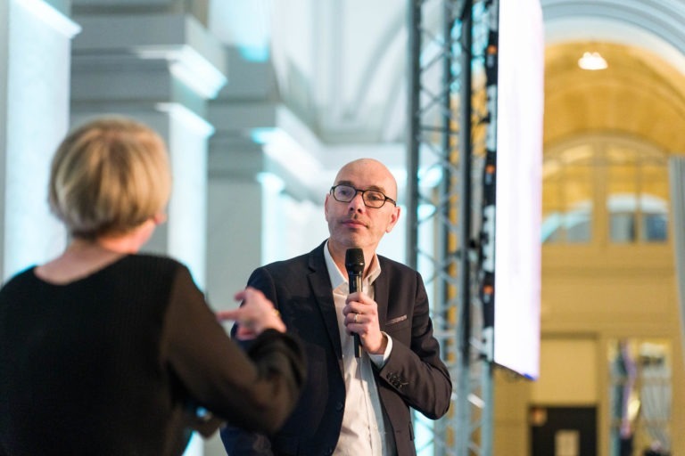 Lors de la Soirée AXA Business au Palais de la Bourse de Bordeaux, un homme en costume et lunettes parle dans un micro tout en regardant une femme aux cheveux blonds courts au premier plan. La scène se déroule à l'intérieur, avec des piliers blancs et des arches en arrière-plan, illuminés par une lumière douce.