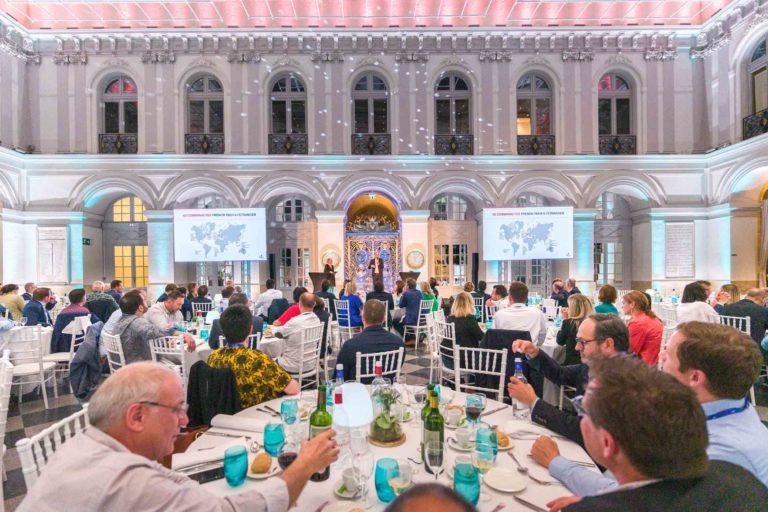 Une grande et élégante salle de banquet du Palais de la Bourse de Bordeaux accueille la Soirée d'affaires AXA. De hauts plafonds et des fenêtres cintrées encadrent des invités habillés de façon formelle, assis à des tables rondes avec des nappes blanches. Des écrans de projection affichant des cartes et des motifs muraux complexes sont mis en valeur par un éclairage rose doux.