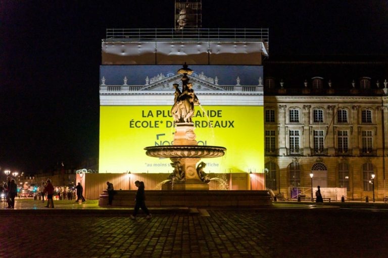 Scène nocturne avec une fontaine illuminée et une statue à plusieurs personnages au centre d'une place. Derrière elle se trouve un bâtiment recouvert d'échafaudages avec une publicité jaune indiquant « LA PLUS GRANDE ÉCOLE* DE BORDEAUX ». Pendant la soirée, AXA Business illumine le Palais de la Bourse tandis que les piétons passent, flanqués de vieux bâtiments.