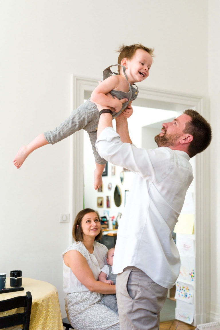 Un homme en chemise blanche soulève un enfant rieur en salopette grise tandis qu'une femme, avec un bébé sur ses genoux, leur sourit depuis la table. La pièce lumineuse, ornée de dessins d'enfants, capture parfaitement la chaleur et la joie de ce moment - une véritable scène de Photographe de famille Bordeaux.