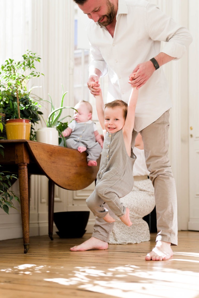 Un homme tient son bambin par les mains tandis que l'enfant se balance avec enthousiasme. Le bambin est pieds nus et vêtu d'une combinaison grise. En arrière-plan, un bébé habillé de la même manière est assis sur une table en bois entourée de plantes en pot avec une douce lumière naturelle qui traverse les fenêtres - parfait pour Photographe de famille Bordeaux.