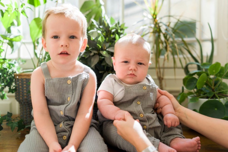 Deux jeunes enfants assis à l'intérieur, entourés de plantes vertes et de soleil. L'enfant le plus âgé, aux cheveux blonds courts, est à gauche, portant une salopette grise avec des boutons en bois. Le plus jeune bébé, également en salopette grise, a une expression sérieuse tout en étant soutenu par les mains d'un adulte. Capture parfaite par le photographe de famille Bordeaux.