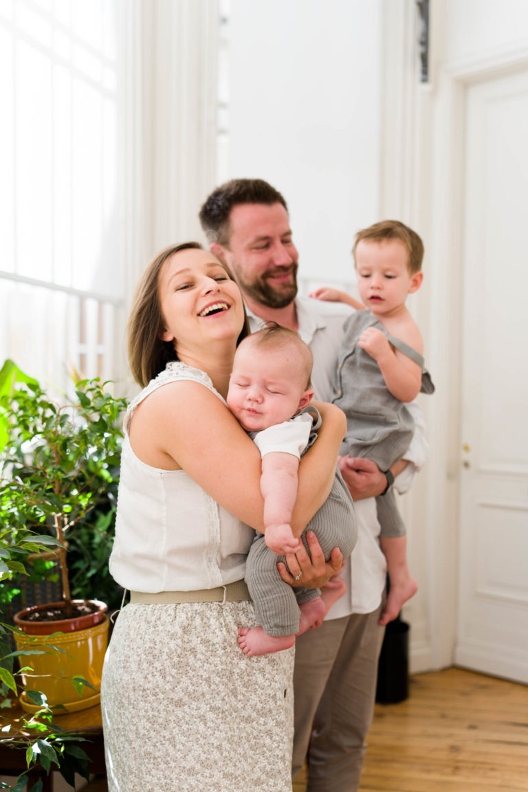 Une femme souriante tient un bébé endormi, debout à côté d'un homme barbu qui tient un petit enfant dans ses bras. La famille, photographiée par un photographe de famille Bordeaux, se tient dans une pièce lumineuse aux murs blancs et aux grandes fenêtres, entourée de plantes vertes en pot. Les adultes sont habillés de vêtements décontractés de couleur claire, et les enfants de tenues grises.