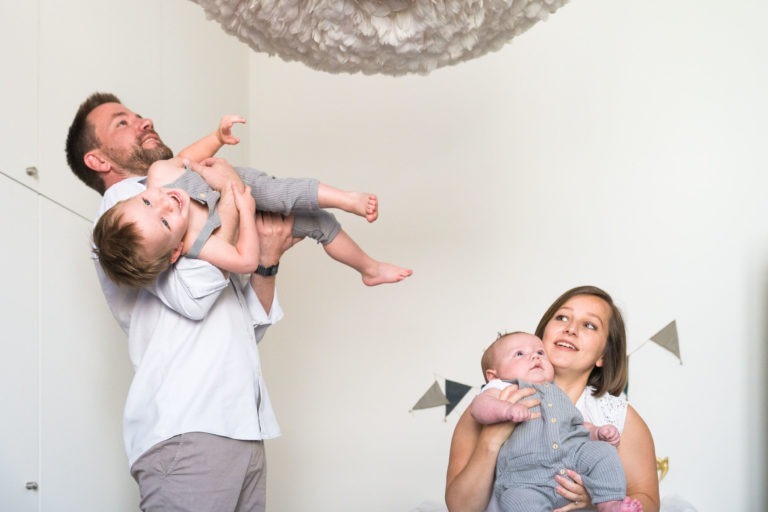 Un homme et une femme jouent avec deux bébés dans une pièce lumineuse. L'homme soulève un bébé très haut dans les airs, le faisant rire, tandis que la femme tient l'autre bébé près d'elle, souriant. Les deux bébés sont habillés de tenues grises assorties. La pièce, photographiée par un photographe de famille Bordeaux, est décorée de fanions et d'un luminaire moelleux.