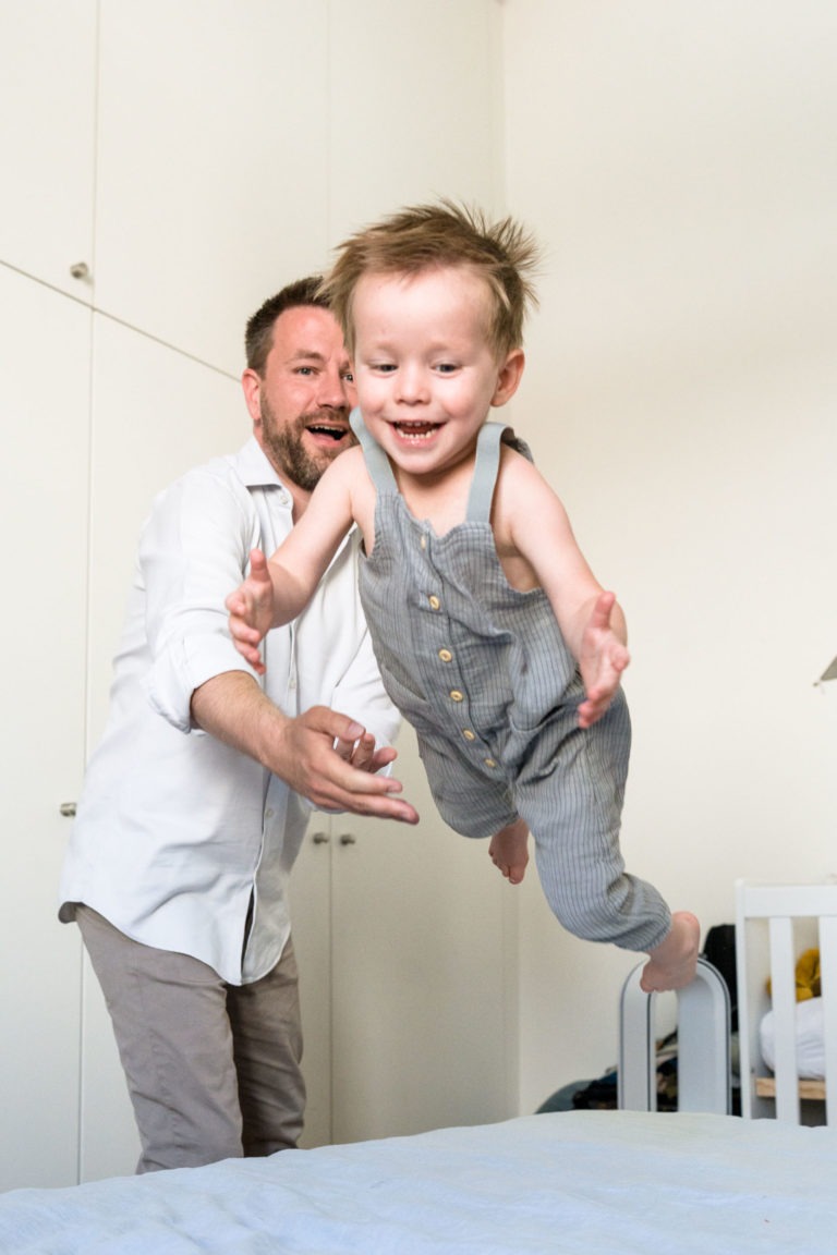 Un homme joyeux avec une barbe et une moustache, vêtu d'une chemise blanche et d'un pantalon beige, attrape un jeune enfant souriant qui est en plein vol. L'enfant porte une combinaison grise sans manches. Ils se trouvent dans une pièce de couleur claire avec des armoires blanches en arrière-plan, parfaitement capturée par le photographe de famille Bordeaux.